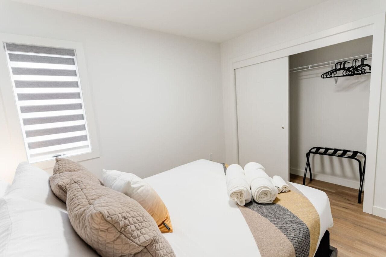 A neatly made bed with a beige quilt, a white comforter, and a striped blanket. There are rolled towels on the bed. A black metal chair is visible in the corner.