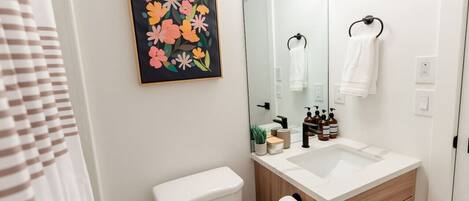 Un baño moderno con inodoro, lavabo y espejo. Hay una obra de arte floral enmarcada en la pared, un perchero y artículos de tocador sobre el lavabo.