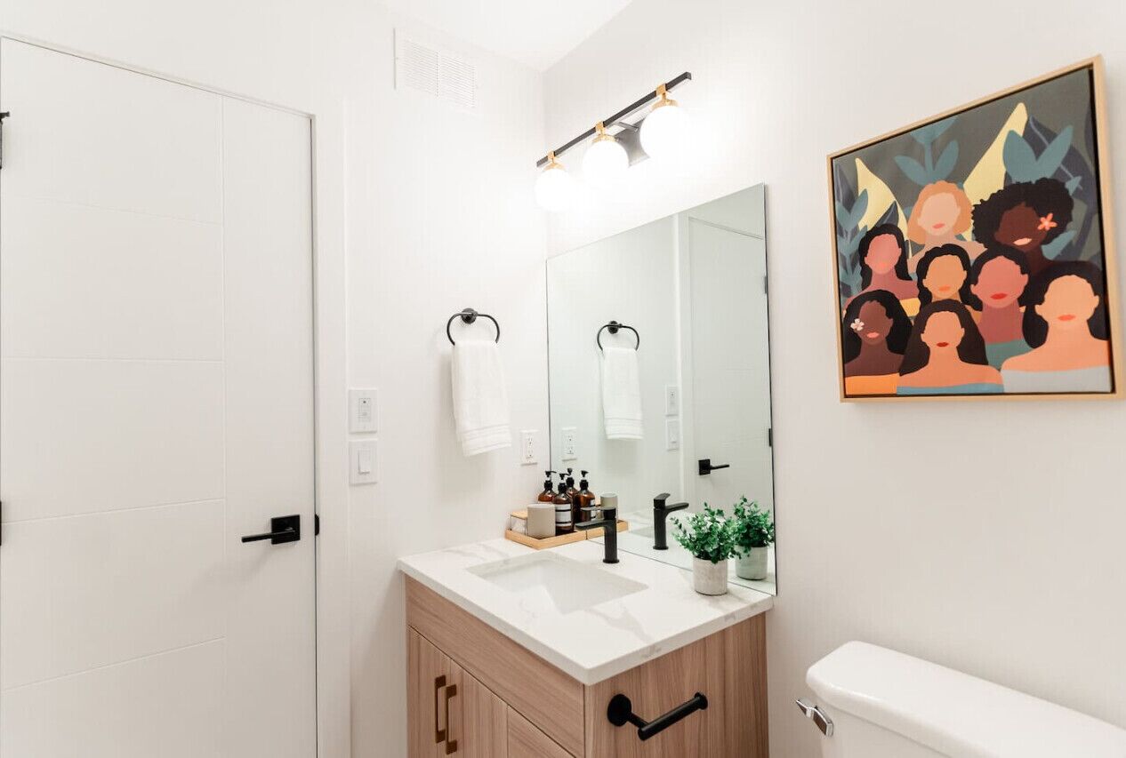 A modern bathroom with a white vanity, a large mirror, and a toilet. There is a framed artwork on the wall above the mirror.