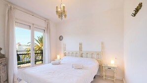 A large, white, double bed with two bedside tables, a chandelier, and a balcony with a view of palm trees. - Costa Brava X-3 in Calella de Palafrugell (Palafrugell)