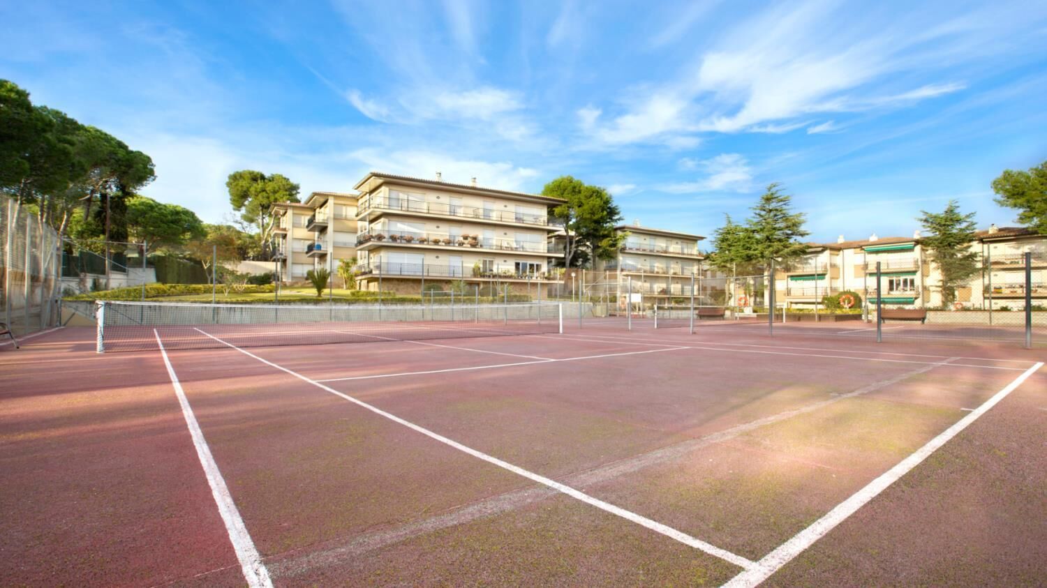An outdoor tennis court with a net, surrounded by buildings and trees.