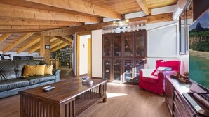 Ein gemütliches Wohnzimmer mit Holzbalkendecke, einem Ledersofa, einem roten Sessel und einem großen Fenster mit Bergblick.