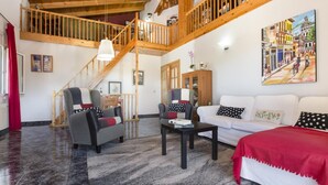 Ein großzügiger Wohnbereich mit hoher Holzbalkendecke, einer Treppe, einem weißen Sofa, zwei Sesseln, einem Couchtisch und einem Gemälde an der Wand.