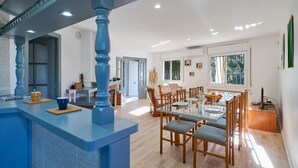 Un vaste espace de salle à manger avec une grande table et des chaises, un coin cuisine avec un escalier bleu, et une vue sur l’extérieur grâce à de grandes fenêtres.