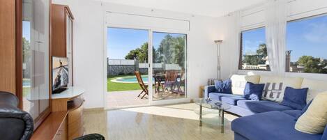 A spacious living room with a blue sofa, a black leather chair, and a glass coffee table. There is a sliding glass door leading to a patio with a pool and outdoor furniture.