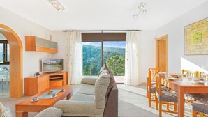 Un salon moderne comprenant un canapé, un téléviseur, une table à manger et des chaises. Une grande fenêtre offre une vue panoramique sur les collines.