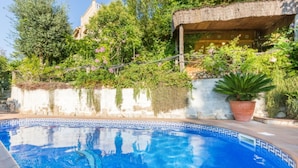 Une piscine bordée de mosaïques bleues, entourée d’une végétation luxuriante et d’un bâtiment rustique doté d’un toit de chaume.