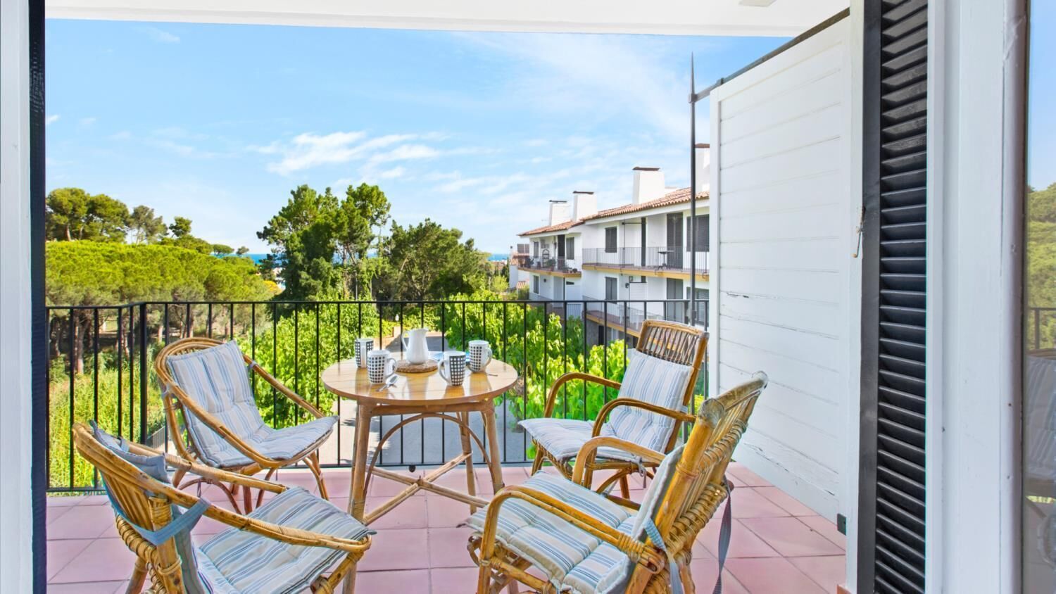 A balcony with wicker chairs and a table, overlooking a residential area with trees and buildings.