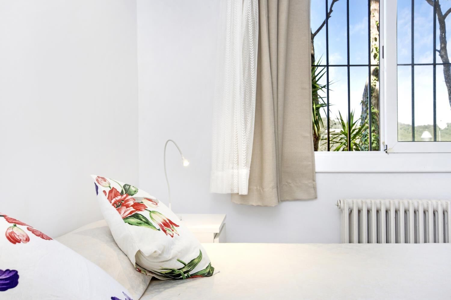 A bedroom with a bed, a nightstand, a window with curtains, a radiator, and a view of greenery outside.