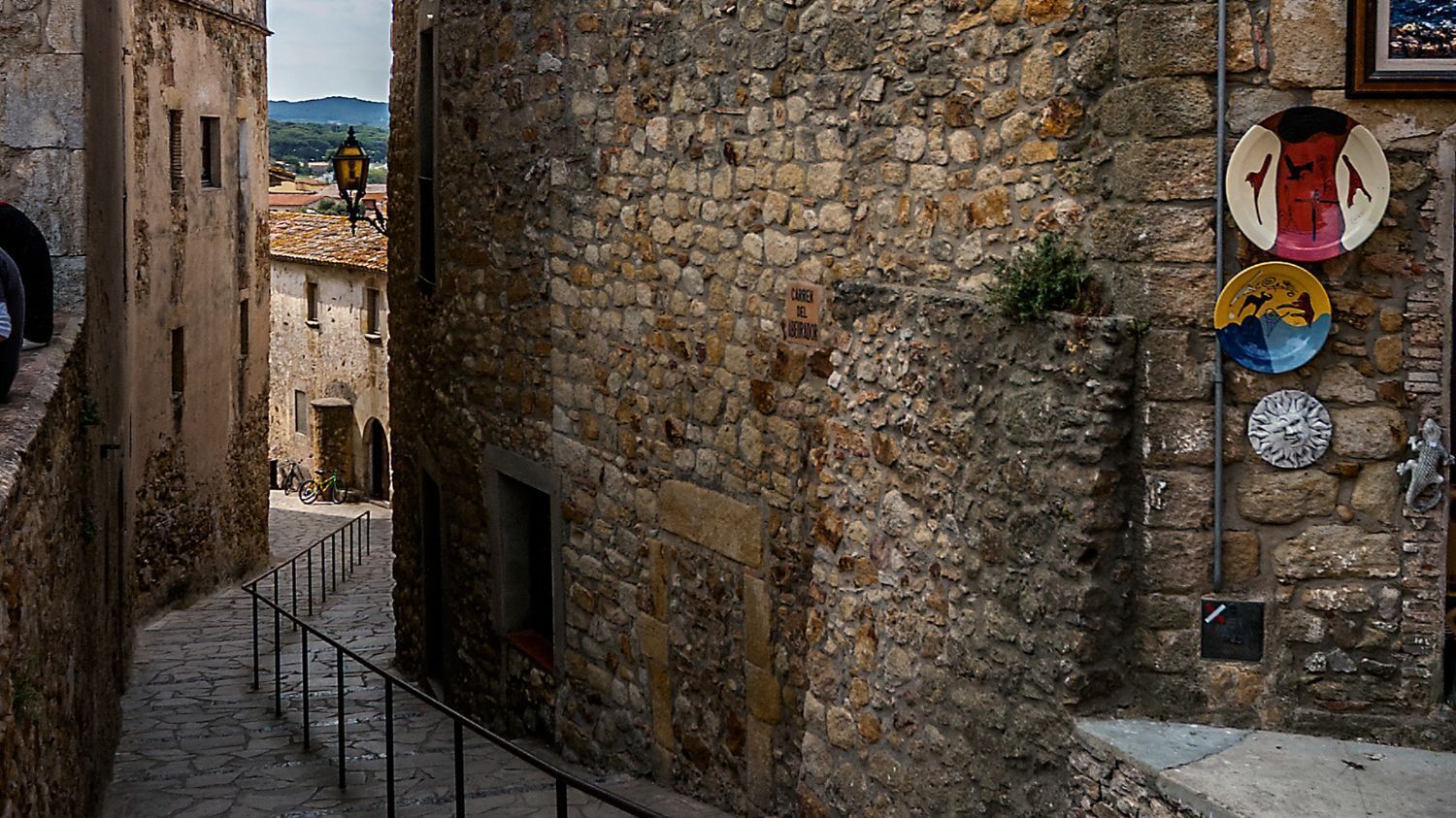 A narrow stone alleyway with a metal railing, a bicycle, and a building with a stone wall and a sign.