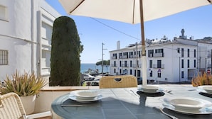 Area esterna con zona pranzo, un tavolo apparecchiato per due, con vista su una strada costiera, edifici e il mare.