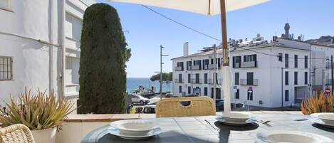 An outdoor dining area with a table set for two, overlooking a coastal street with buildings and a view of the sea.