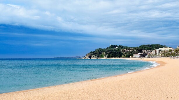 A beach with clear blue water, sandy shore, and buildings on a hill in the background.
