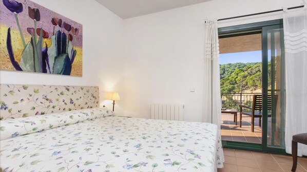 A bedroom with a bed, a painting, a lamp, and a view of a balcony with chairs.