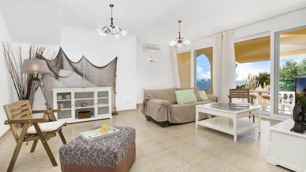 Un salon spacieux avec un canapé, un pouf et une table basse en verre. On y trouve un meuble blanc aux portes vitrées et un lustre suspendu au plafond.