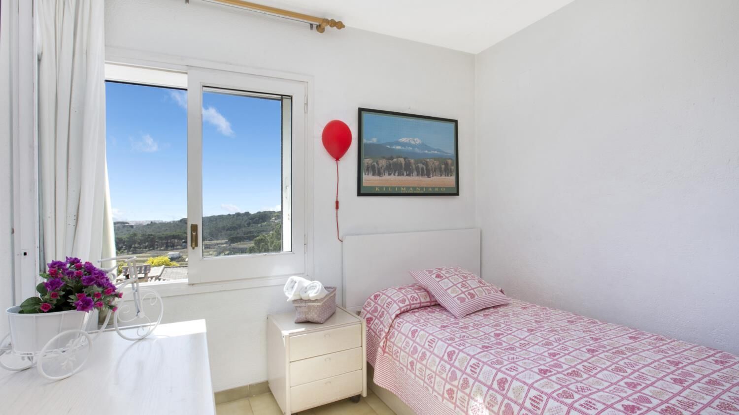 A bedroom with a bed, a nightstand, a window with a view, a framed picture on the wall, and a red balloon.