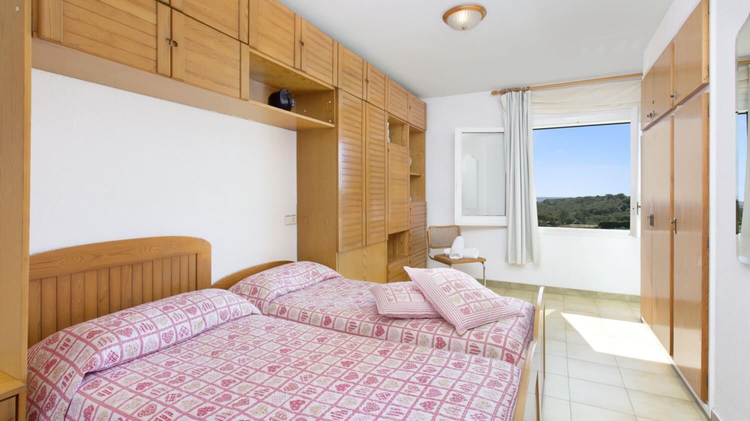 Un dormitorio con dos camas, armarios de madera, una ventana con vistas y una mesita de noche con lámpara.