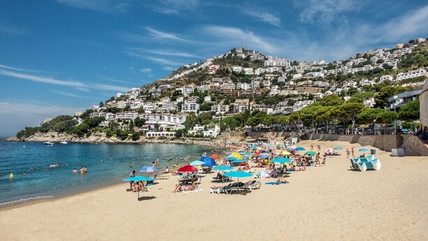 A beach with people swimming and sunbathing, a rocky hillside in the background, and a clear blue sky. - Vila Lina in Lloret de Mar (Lloret de Mar)