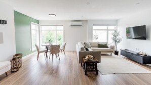 Una sala de estar moderna con un sofá, una mesa de comedor con sillas, un televisor en la pared y un ventanal que proporciona luz natural.