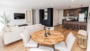 Una sala de estar moderna con una mesa redonda de madera, un sofá blanco y una zona de cocina con armarios oscuros y un taburete de bar.