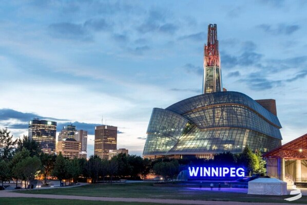 Un edificio moderno con fachada de vidrio curva y la palabra 'WINNIPEG' iluminada en el suelo.