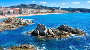 A coastal cityscape with a beach, rocky outcrops, and a row of multi-story buildings.