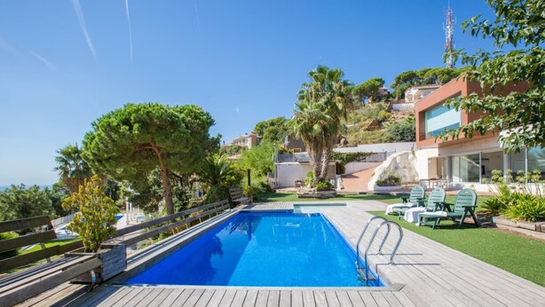 A swimming pool with a wooden deck, surrounded by greenery and a hillside with buildings.