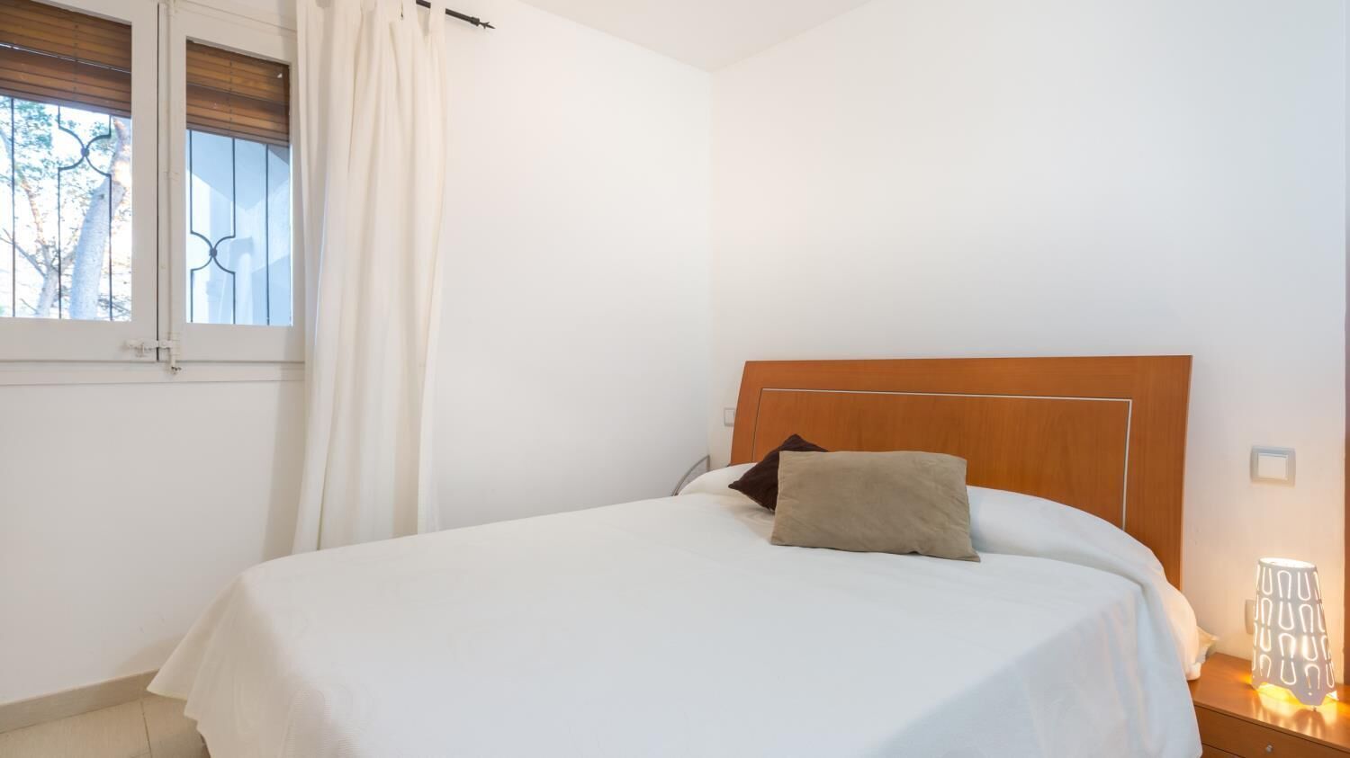 A neatly made bed with a wooden headboard, a bedside lamp, and a window with curtains.