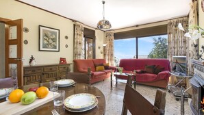 Una sala de estar con chimenea, una mesa de comedor con fruta y platos, un sofá y un ventanal con cortinas.