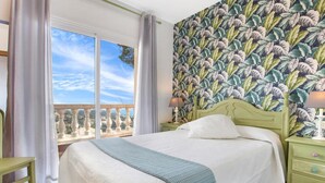 Un dormitorio con un ventanal grande, una cama con colcha blanca, una mesita de noche verde y un papel tapiz floral.