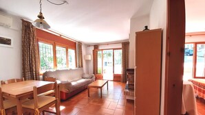 Un salon chaleureux avec un canapé, une table à manger en bois et des chaises. De grandes fenêtres sont agrémentées de rideaux, et on y trouve également un ventilateur de plafond et une porte donnant sur l’extérieur.