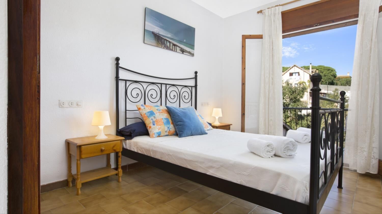 Een slaapkamer met een bed, bedlampjes, een houten nachtkastje, uitzicht naar buiten en een schilderij aan de muur.