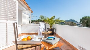 Un balcon avec un fauteuil en osier, une petite table avec une plante, un plateau avec des oranges et un livre, et une vue sur une colline parsemée de bâtiments.