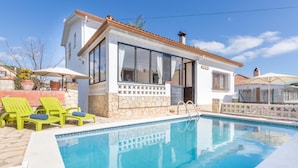 Un espace aménagé au bord d’une piscine à l’eau d’un bleu limpide, un bâtiment blanc avec un toit recouvert de tuiles et des chaises longues vertes.