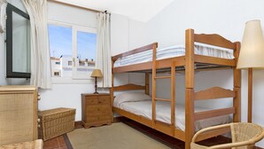 Une chambre avec un lit superposé doté d’une tête de lit en bois, une table de chevet, une lampe, une chaise en osier et une fenêtre avec des rideaux.