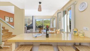 A spacious kitchen with a large wooden table, a wine bottle, and a book. There are multiple windows providing a view of the outdoors. - Ronda in Llafranc With 4 Bedrooms and 3 Bathrooms (Palafrugell)
