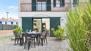 Un edificio de dos pisos con un balcón, un comedor con mesa y sillas, y un pequeño jardín con plantas.