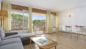 A spacious living area with a sofa, a glass-top coffee table, and a dining table with chairs. Large windows offer a view of trees and a balcony. - Illa 23 in Calella de Palafrugell (Palafrugell)