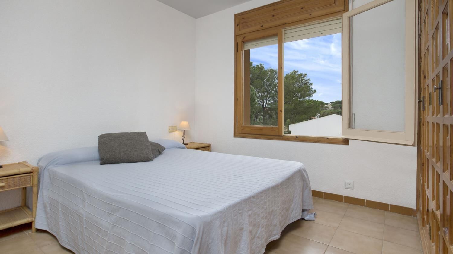 A bedroom with a single bed, a nightstand with a lamp, a window with a view of trees, and a wooden door.