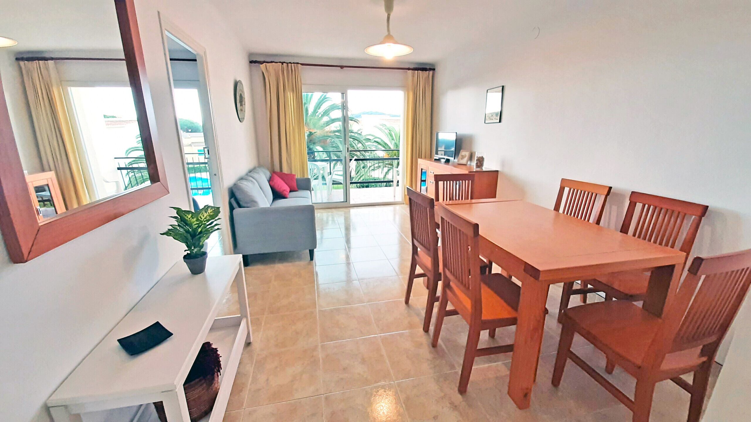 Un comedor con mesa y sillas de madera, un sofá, una mesita con una planta, y vistas a un balcón con cortinas.