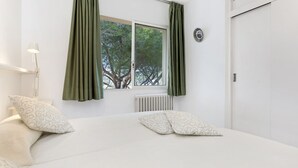 A bedroom with a bed, two pillows, a window with curtains, a radiator, and a wall-mounted light fixture. - Ancora 15 B-2 in Llafranc (Palafrugell)