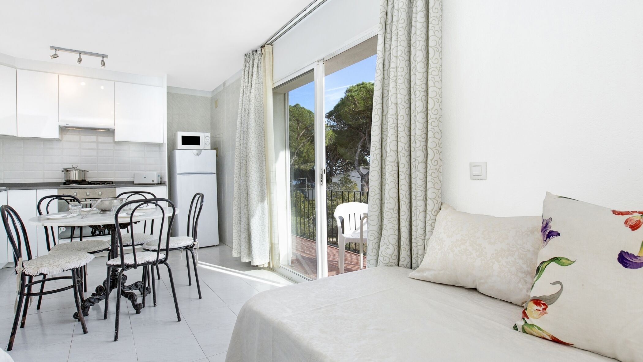 A modern kitchen with white cabinets, a dining table with chairs, a white sofa with floral cushions, and a balcony with curtains.