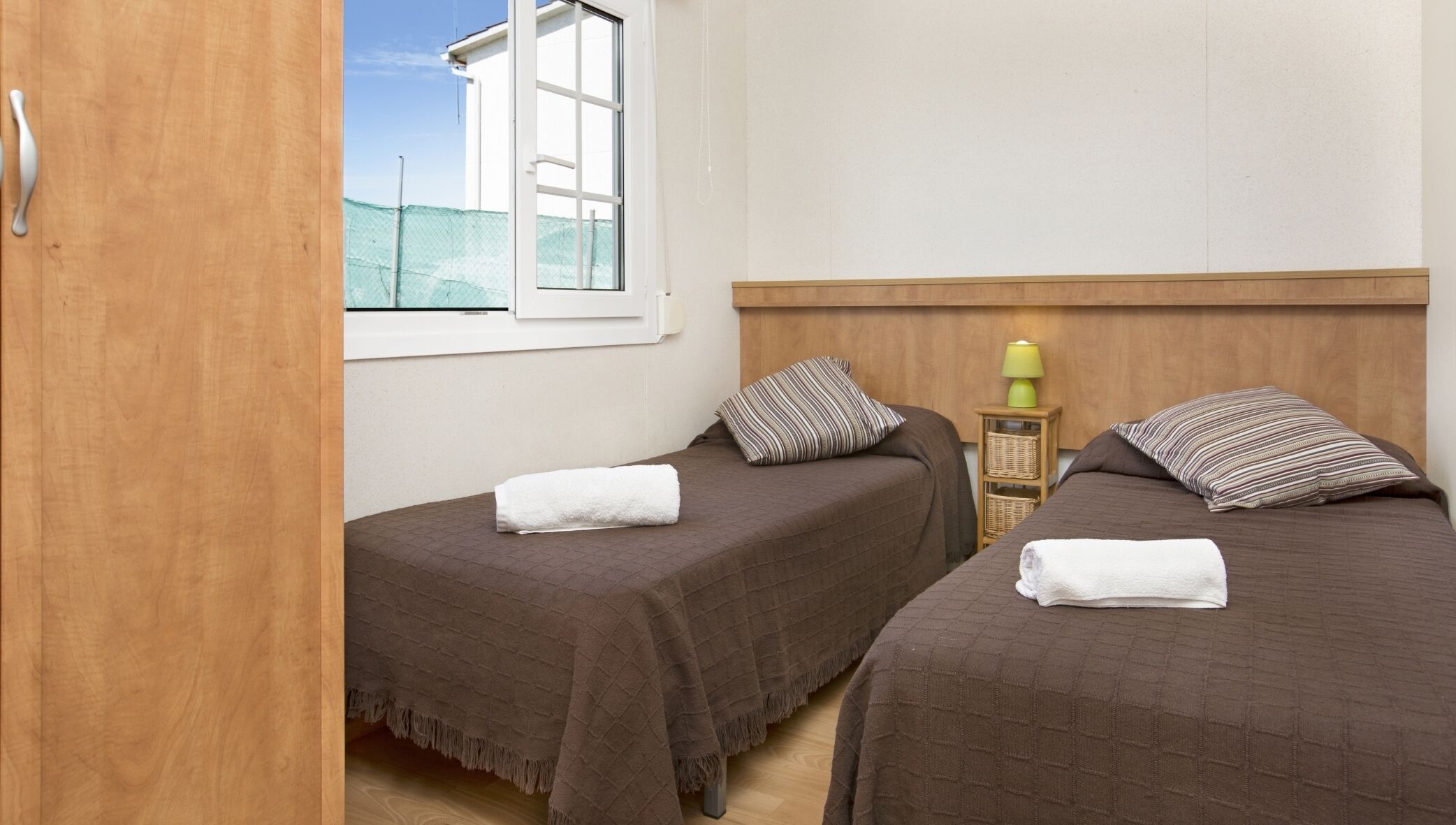 A bedroom with two beds, a window with a view of the outside, a bedside table with a lamp, and a wooden wardrobe.