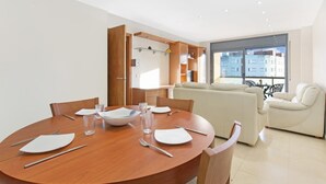 A modern dining area with a round wooden table set for four, surrounded by beige chairs. A sofa and a chair are visible in the background, with a large window providing a view of the cityscape. - Santa Cristina in Lloret de Mar (Lloret de Mar)