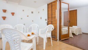 A room with a dining area featuring white plastic chairs and a table, a wooden door, and a bed with a white bedspread. - Pruneda in Llafranc With 2 Bedrooms and 1 Bathrooms (Palafrugell)