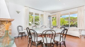 A dining area with a glass table surrounded by chairs, a stone fireplace, and large windows with curtains. - Pandora 1 in Llafranc (Palafrugell)