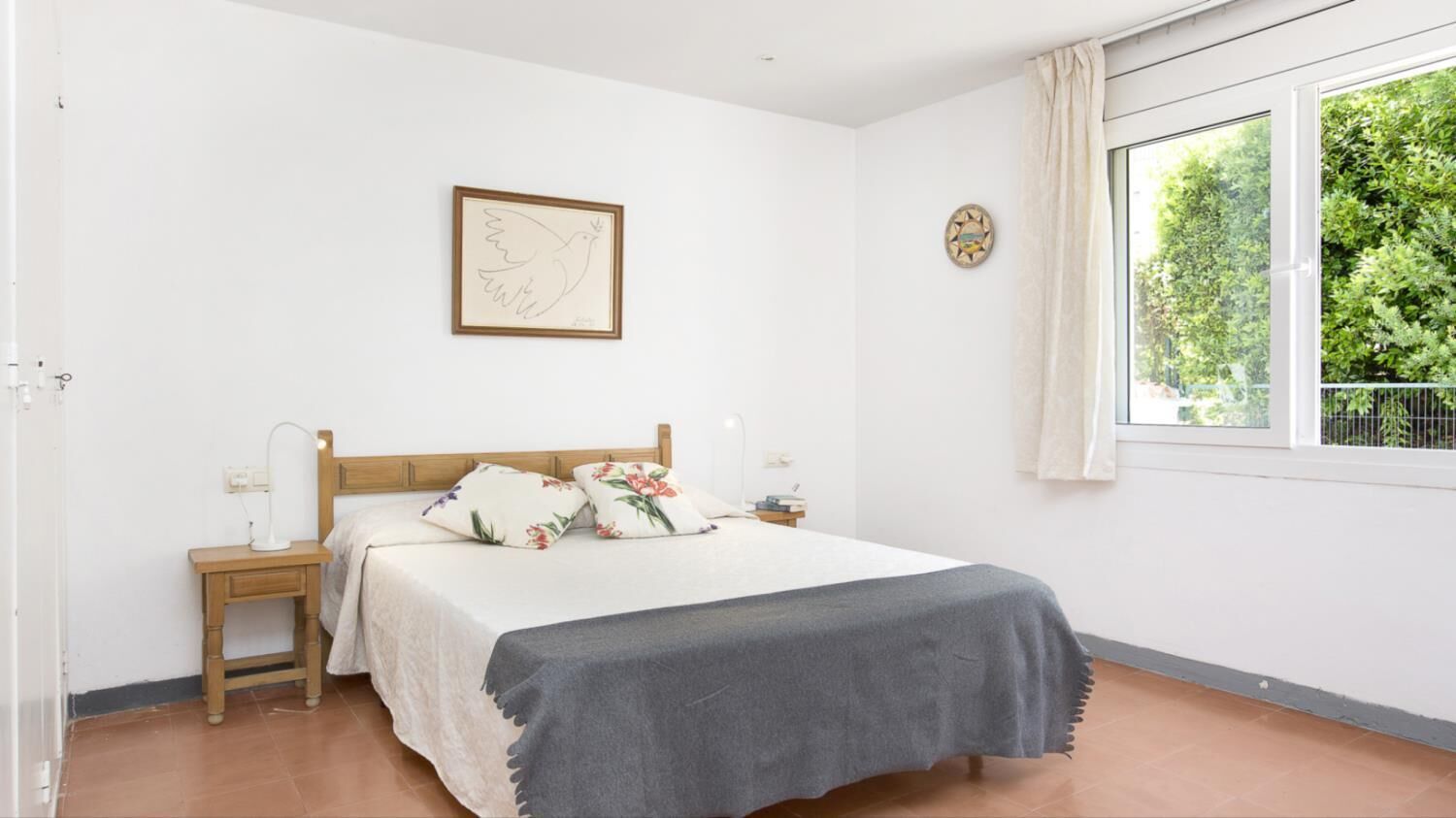 A bedroom with a bed, a nightstand, a lamp, a framed artwork, a clock, and a window with a view of greenery.