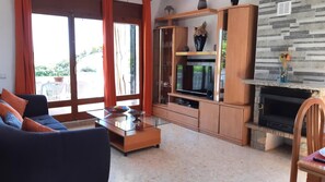 Una sala de estar con sofá, mesa de centro, televisor, chimenea y un balcón con plantas en macetas.