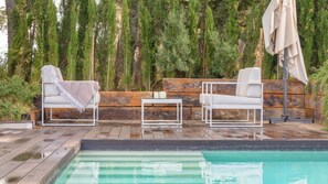 Ein Poolbereich mit zwei weißen Liegestühlen, einem kleinen Tisch und einer Holzterrasse.