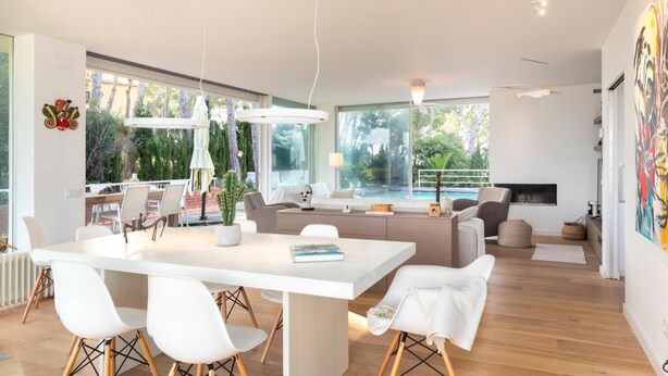 A modern living room with a large white dining table, white chairs, a grey sofa, and a TV. There are large windows with a view of greenery outside. - Ferran in Llafranc With 4 Bedrooms and 2 Bathrooms (Palafrugell)